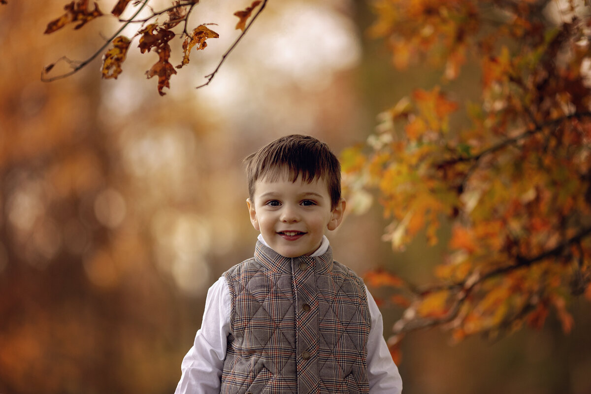 A toddler boy in a brown plaid vest stands in a park at sunset in the fall