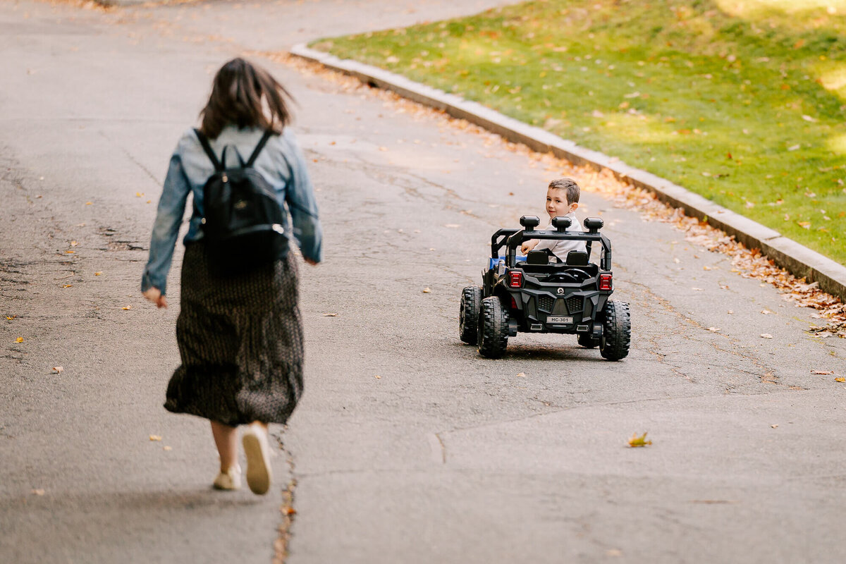 Mother with a backpack on runs down a quiet suburban street, her young son driving his battery-powered car ahead of her, turning his head and laughing.