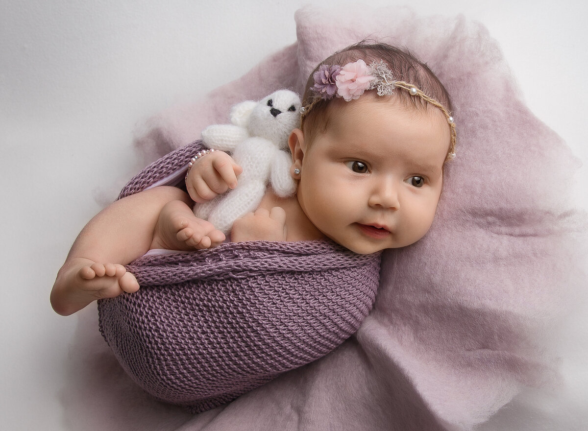 Newborn baby girl is awake for her newborn photoshoot. She is bare and wrapped in a purple blanket. In her hands she is holding a tiny white teddy. Captured by top Brooklyn newborn photographer Chaya Bornstein
