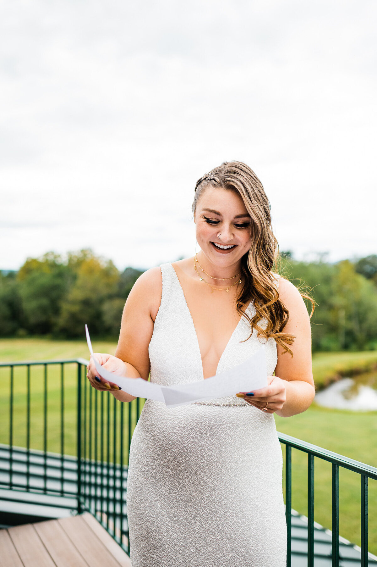 bride opening up a letter from her groom and reading it while standing on balcony with a large open field behind her with a lush tree line