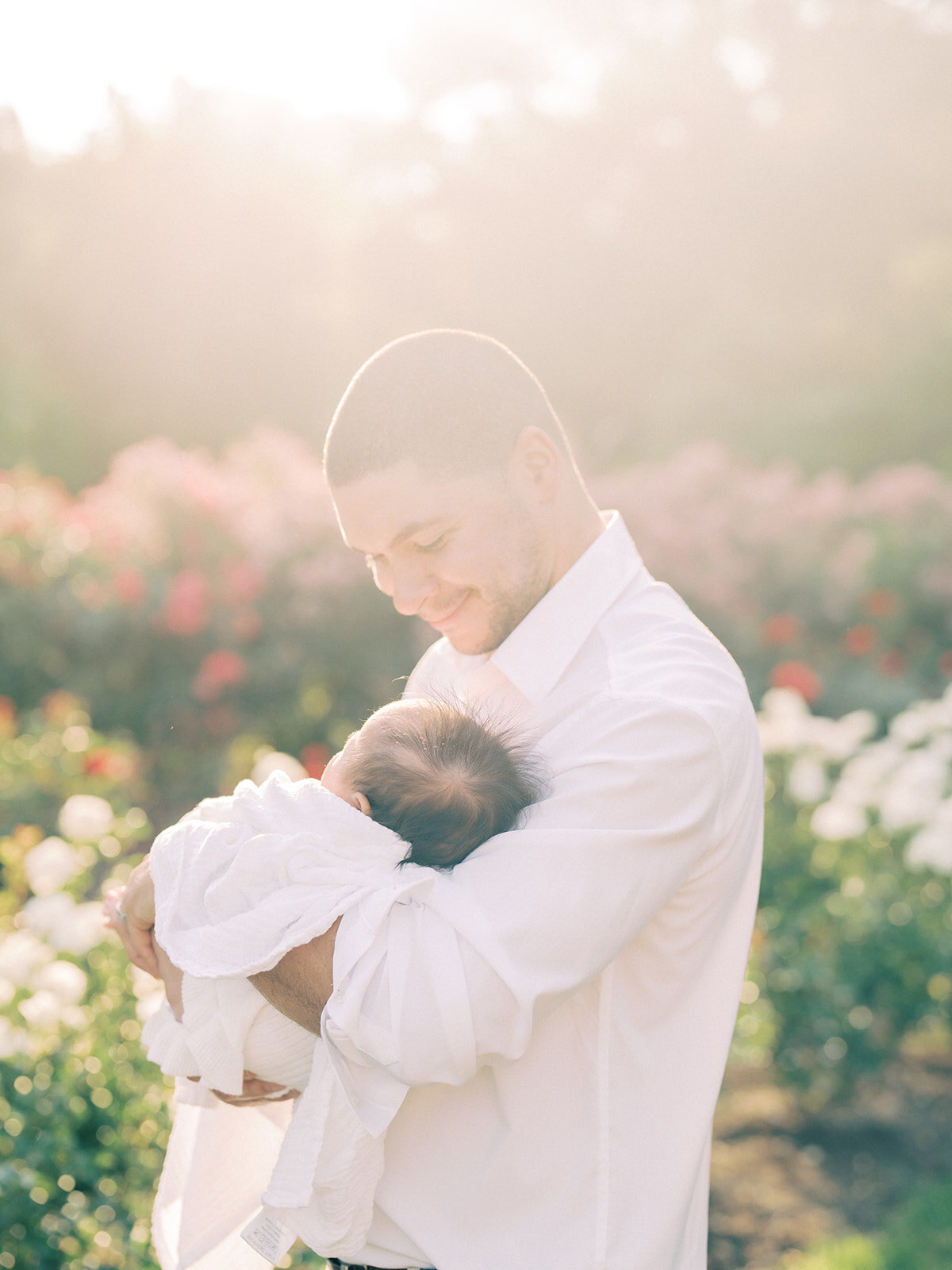 Father smiles down at his newborn baby girl as he holds her in Bon Air Rose Garden at sunset.