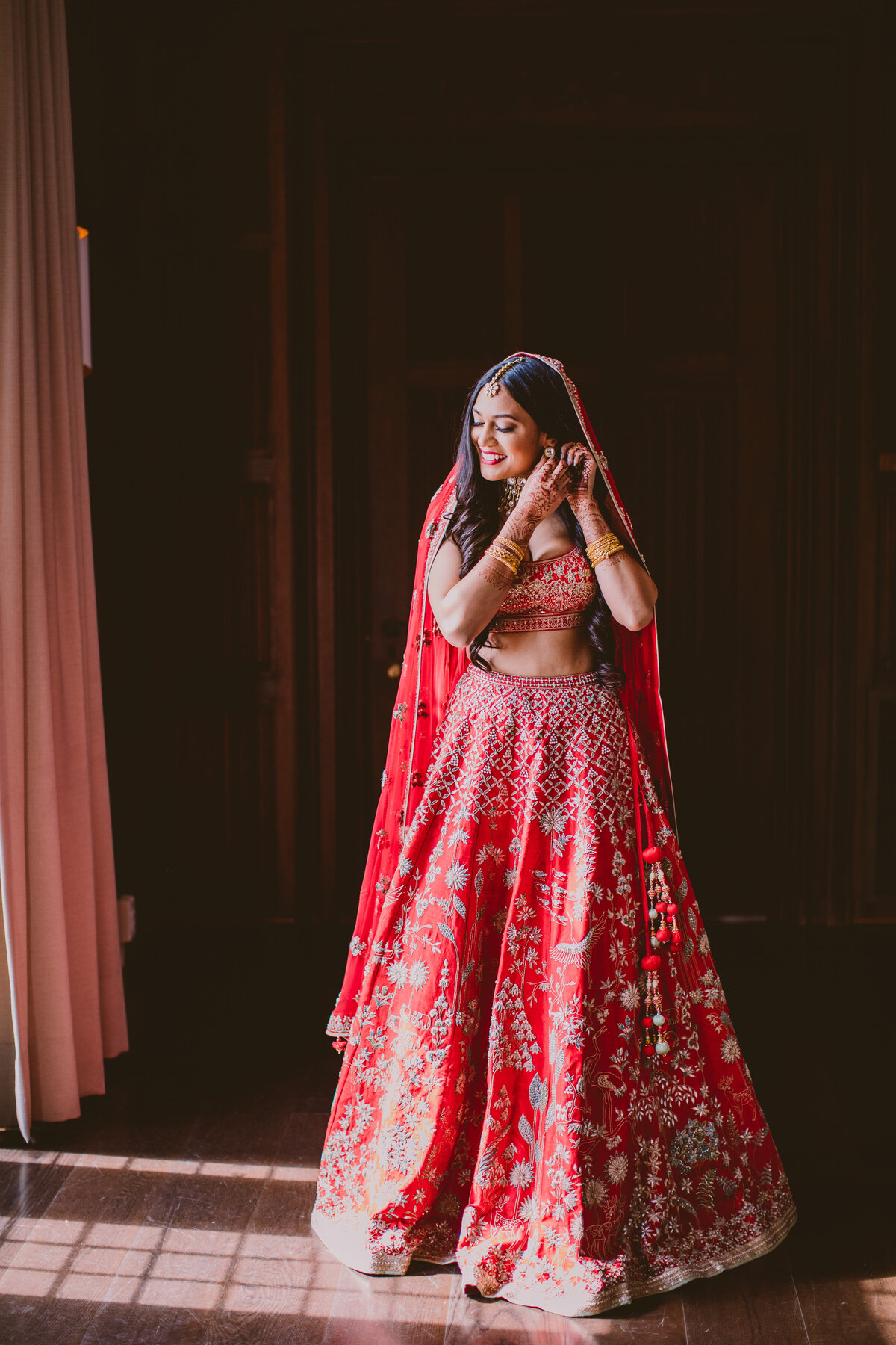 indian bride getting ready during her bridal prep in front of a window wearing a red lehnga inside natirar mansion
