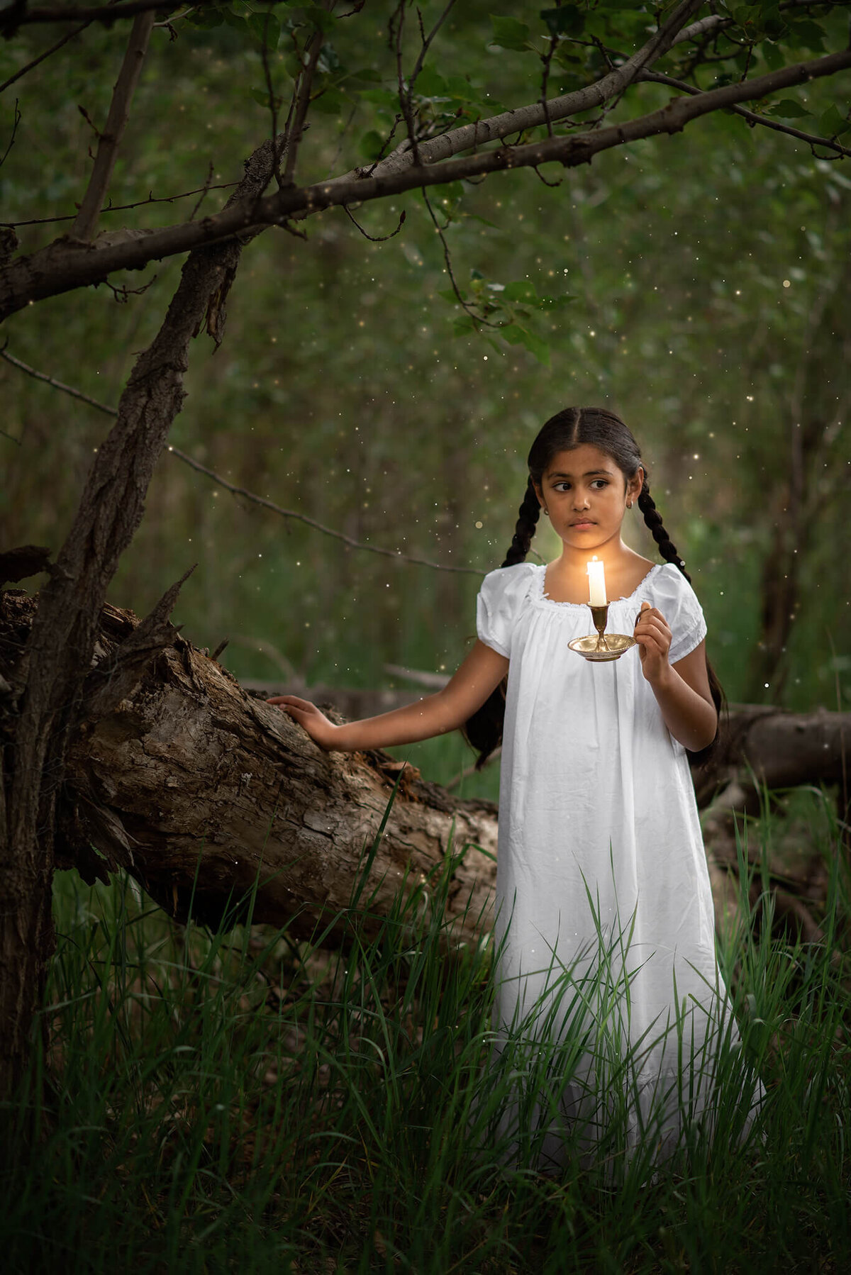 A young girl in a white nightgown holds a candle in the dark forest, surrounded by fireflies, in Kamloops BC. Captured by Brittany Danielle