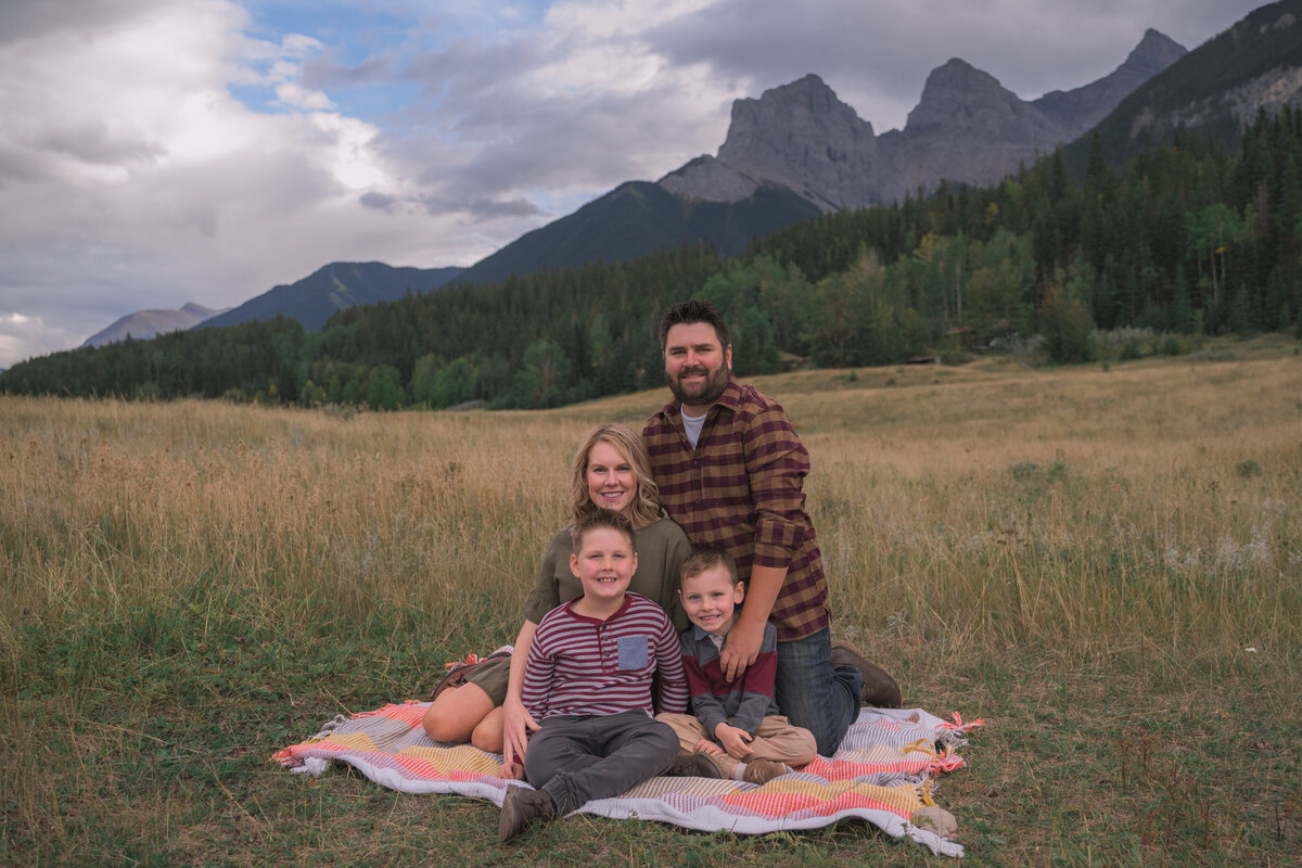 Young family sits on a blanket in grassy meadow near Canmore with mountains behind.