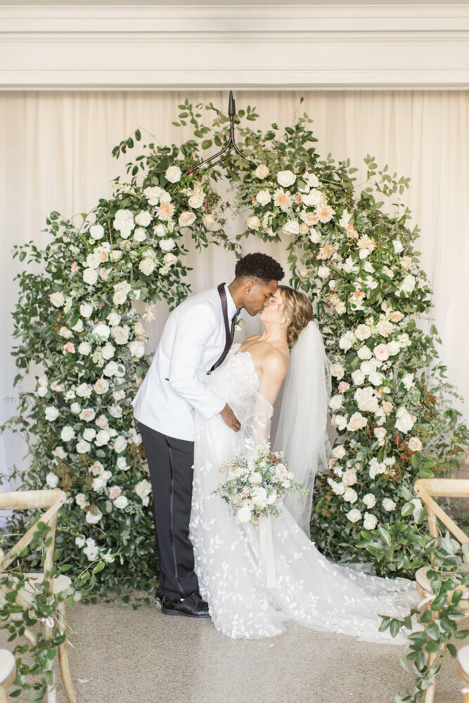 Bride and groom kiss under arch of beautiful flowers at wedding ceremony