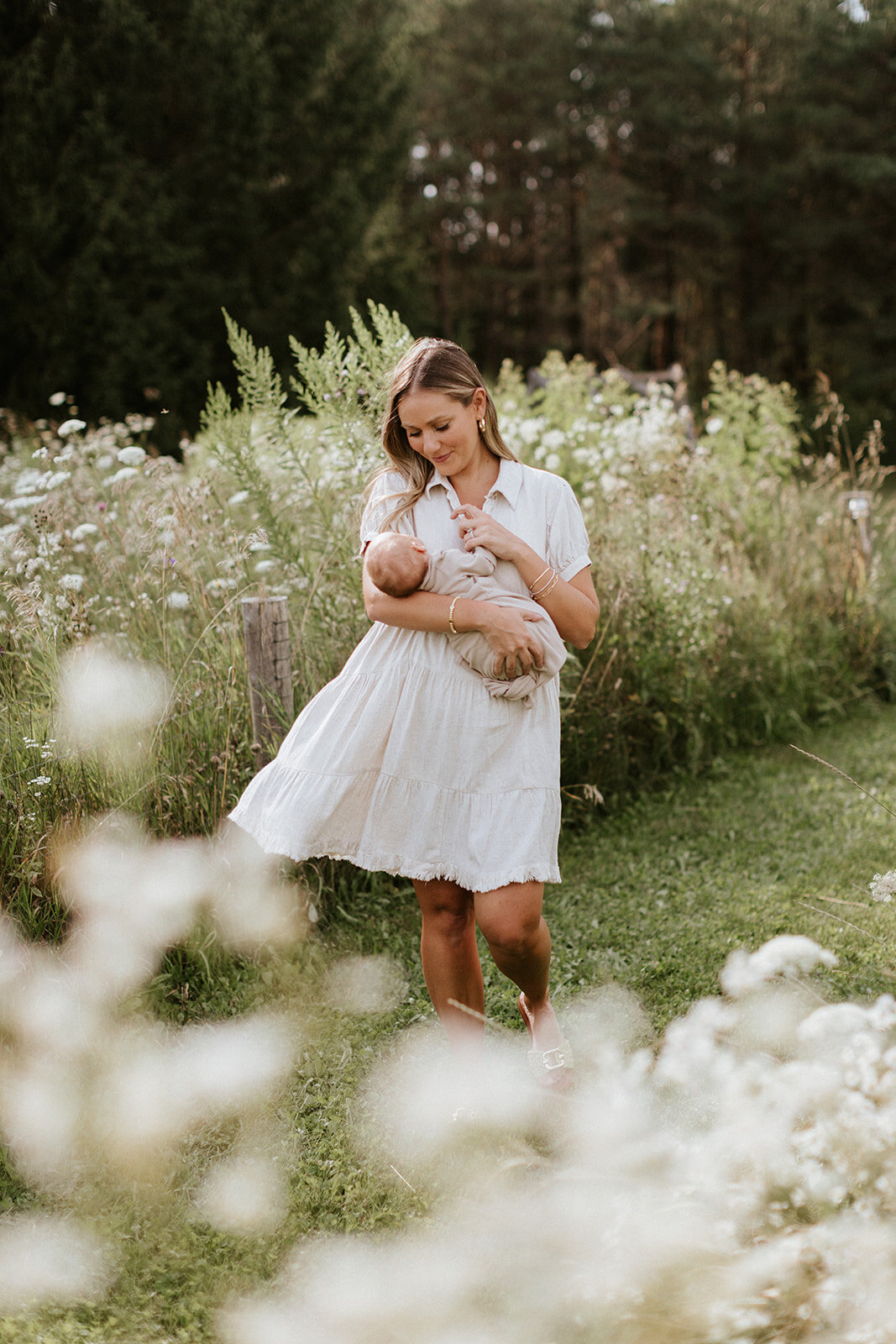 A woman in a white dress holds a baby amidst the wildflowers and greenery, captured by newborn photographer, Jennifer van Son.