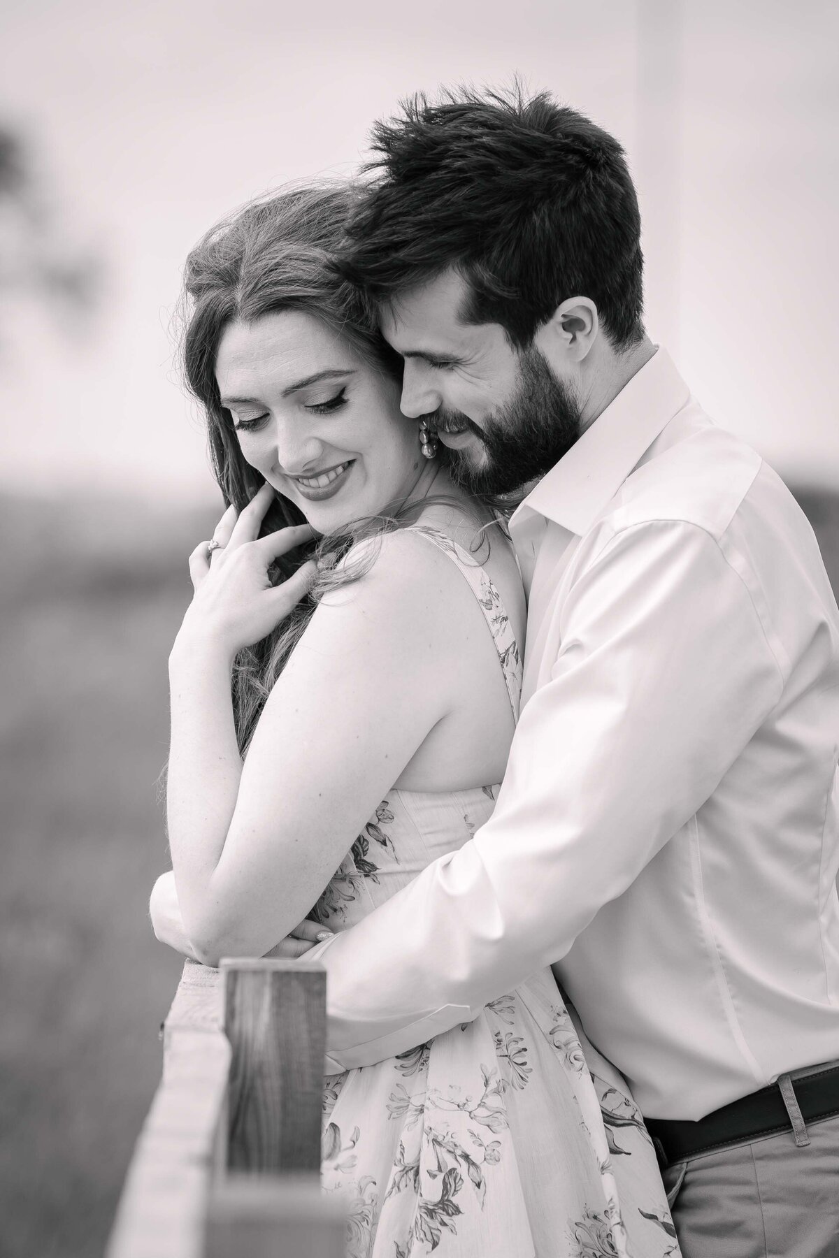 A black and white photo of an engaged couple resting on a fence cuddling