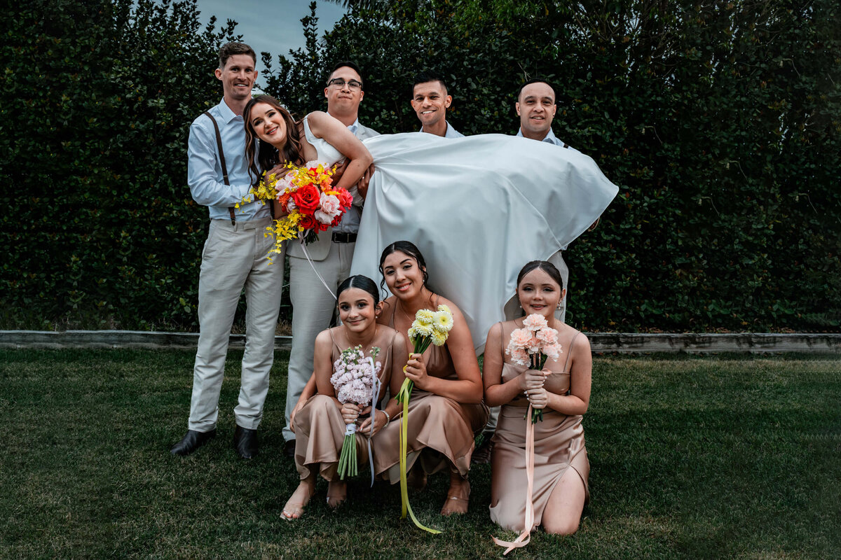 A joyful wedding party stands together on the grass, smiling for a group photo in a beautiful outdoor setting.   - photo by Jamie Simmons