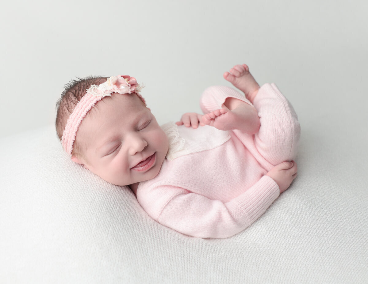 A cute newborn girl adorned with a delicate flower headband, smiling softly in our Rochester, NY photography studio, displaying her charm.