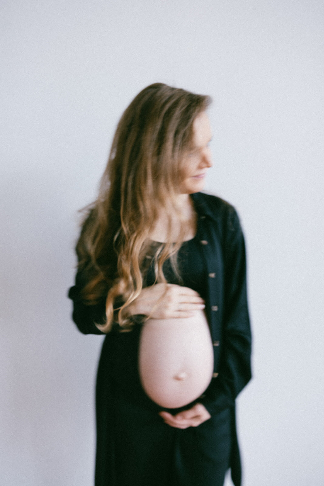 Woman dressed in black holds her belly in the natural light of a studio photo session in Oregon.