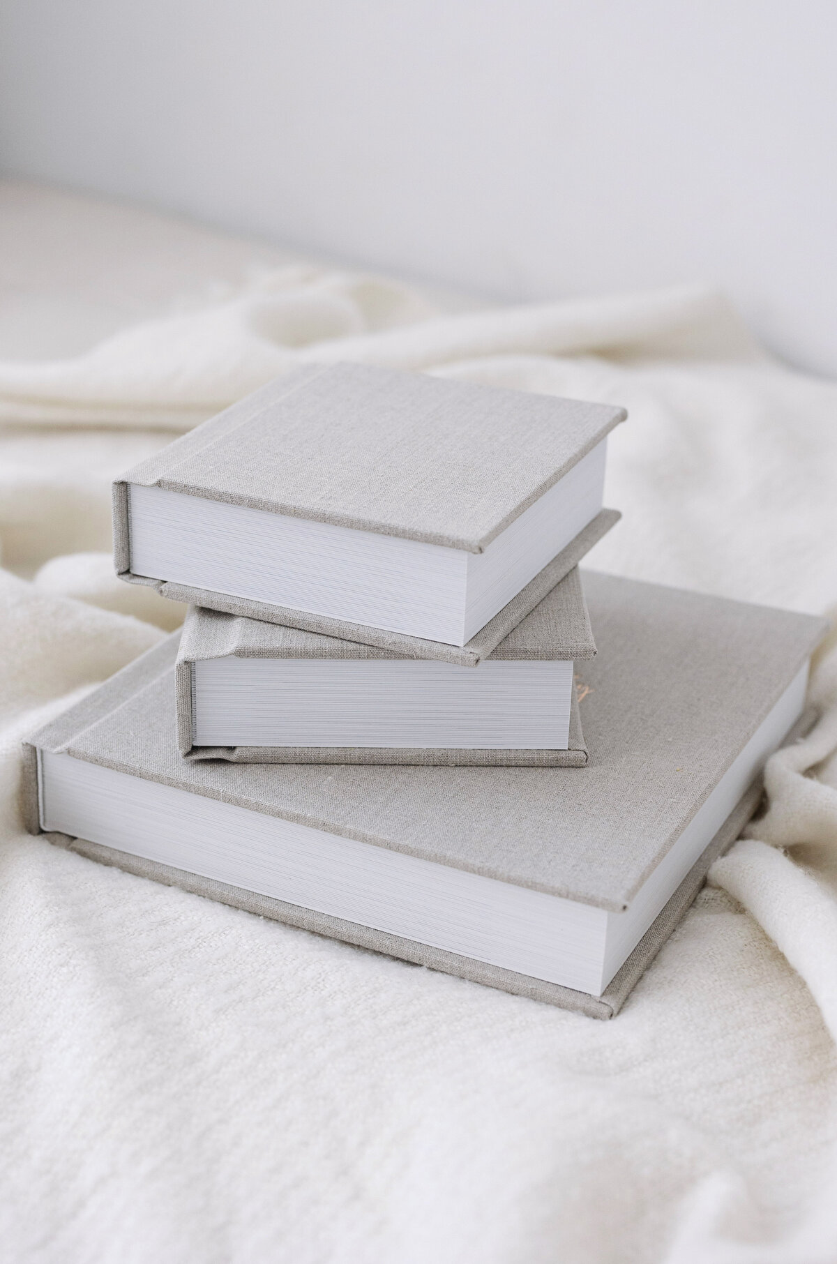 Three gray hardcover books of varying sizes stacked on a soft white fabric surface. The books have a minimalist design, with no visible text or illustrations on the covers. Lighting is soft, creating a calm and cozy atmosphere.