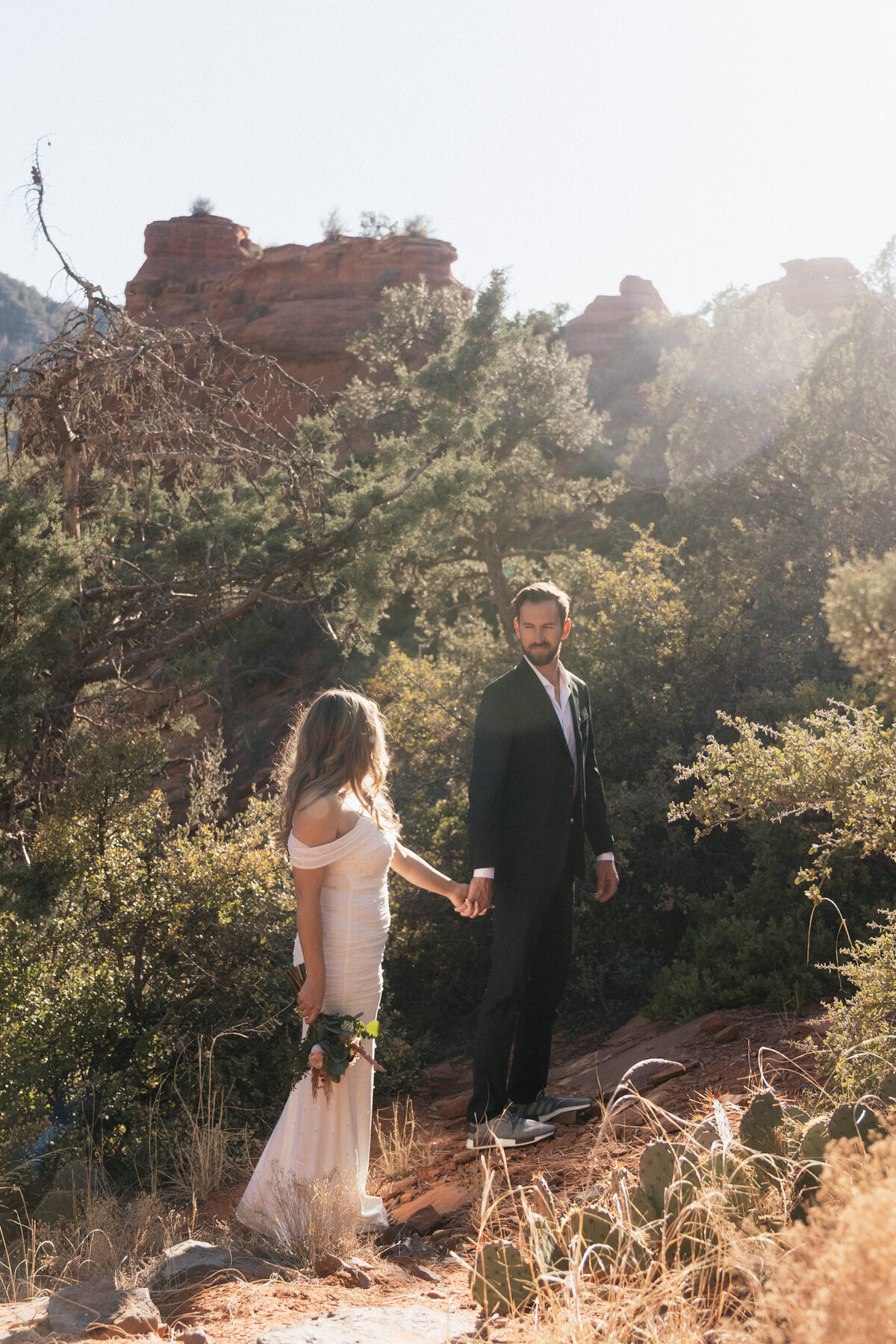 Bride and groom holding hands during their adventurous elopement at Merry Go Round Rock in Sedona, Arizona, surrounded by desert vegetation and red rock formations in golden sunlight.