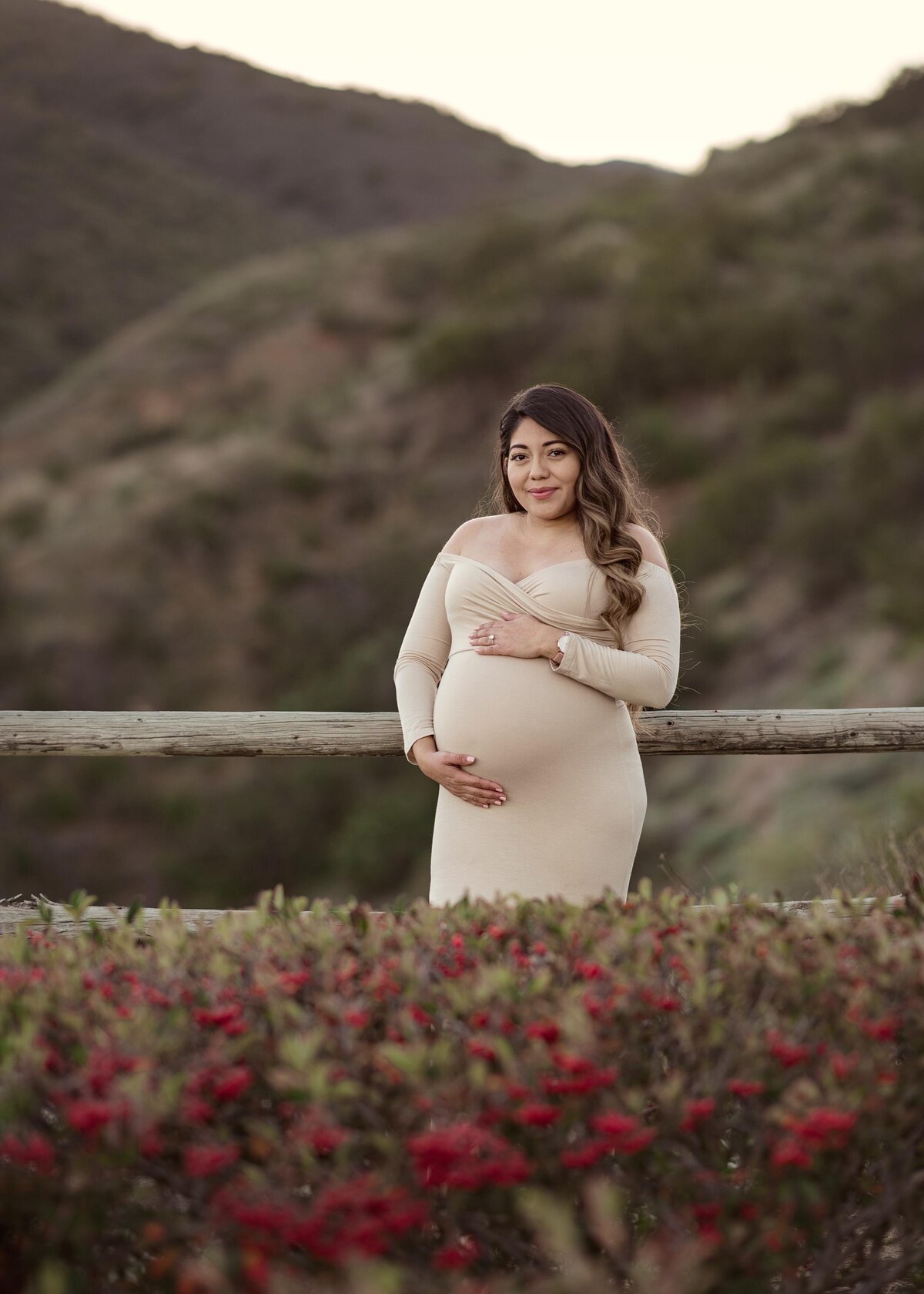 Mom in long, cream, off-the-shoulder maternity gown holding her bump and smiling at the camera. Elsinore mountains in the background, dark red floral bush in the foreground.