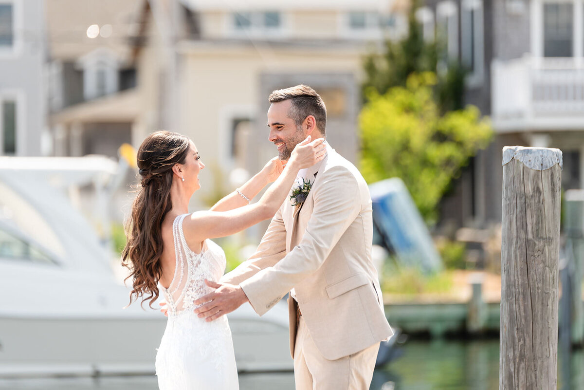A bride and groom share a joyful moment, hugging on a sunny dock with boats and waterfront homes in the background.