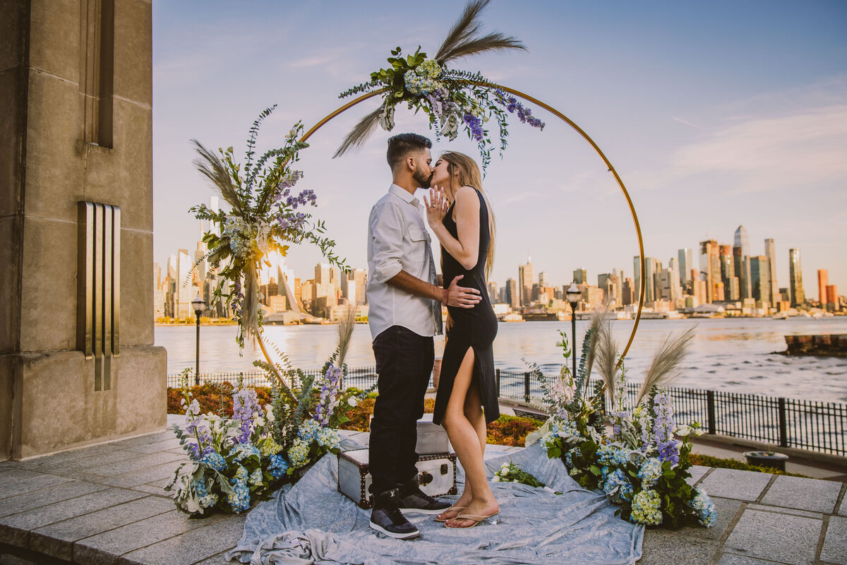 An engaged couple during their proposal posing with the ring and kissing in front of a decorated arch at the riverwalk view in west new york in new jersey