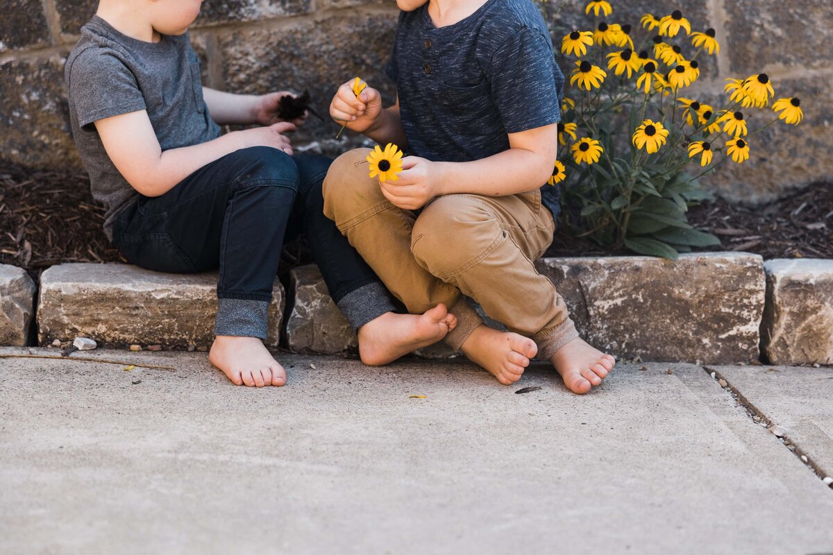 Two children sitting on a curb, one holding a yellow flower; barefoot with black-eyed susans in the background. Captured by a family photographer in Pittsburgh, PA.