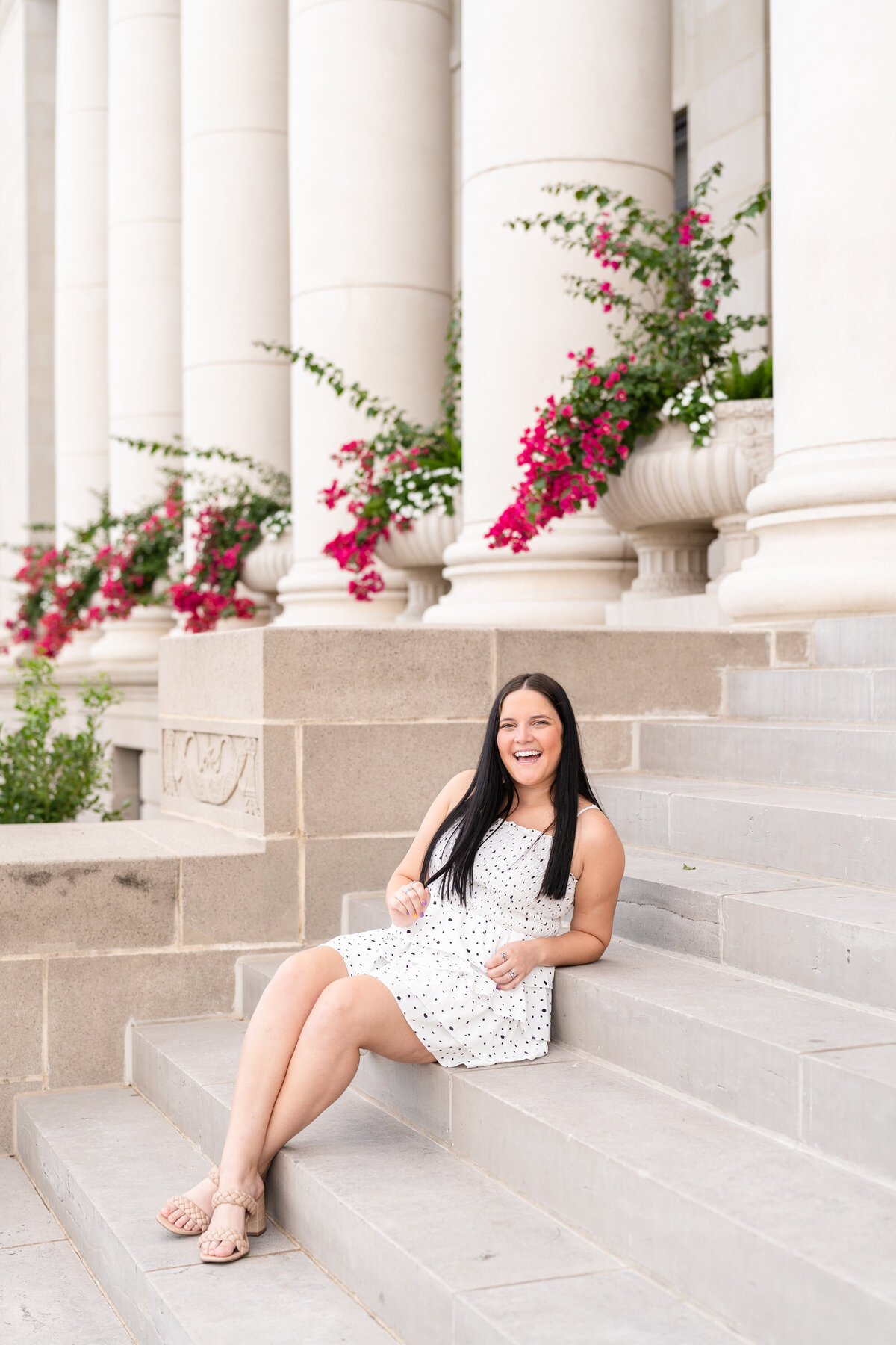 Texas A&M senior girl sitting on Admin Building stairs laughing at camera while wearing white dress with pink flowers in background