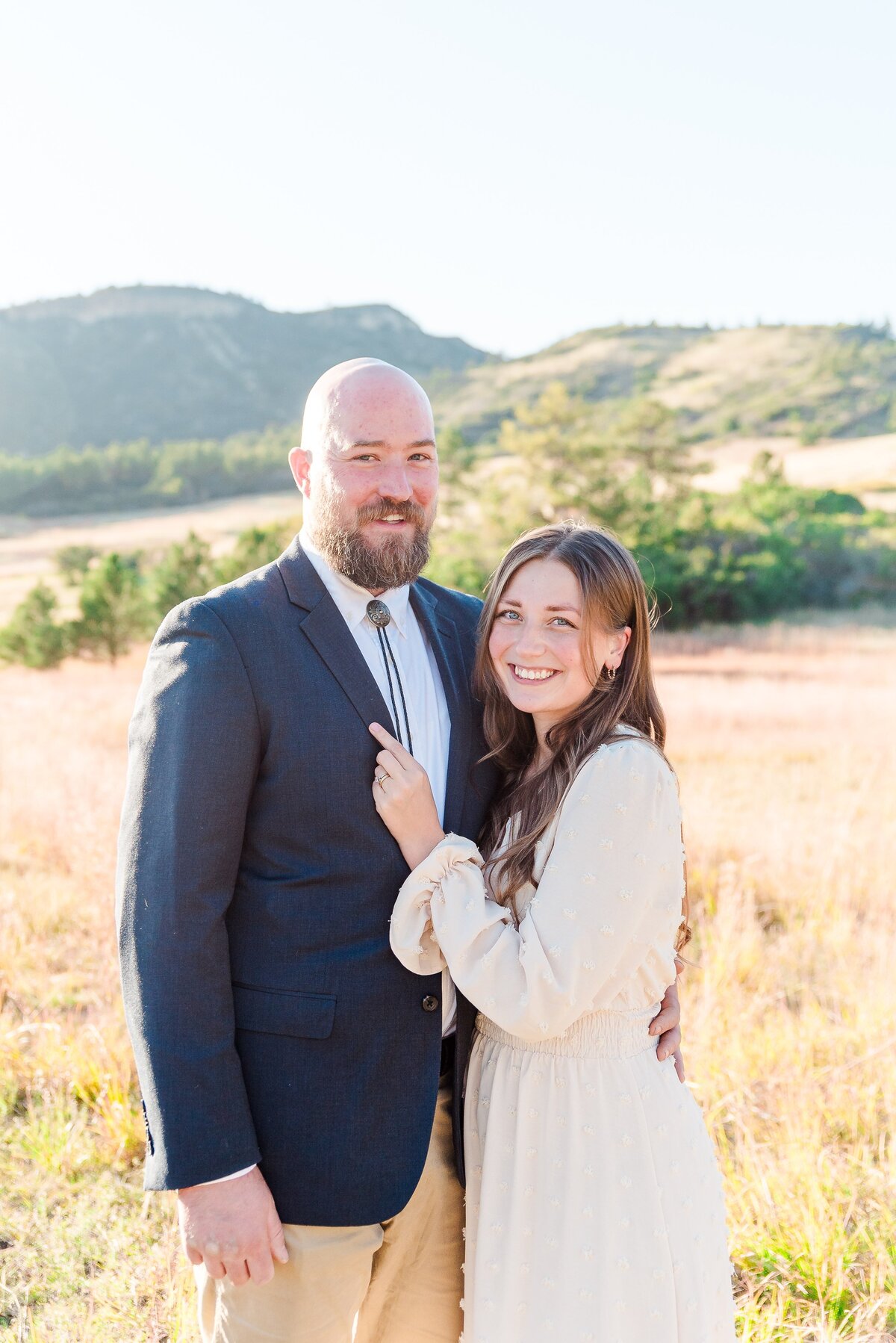 a woman in a tan dress with long sleeves and long brown hair holds on to her partners blue jacket as they both smile for the camera captured by denver family photographer