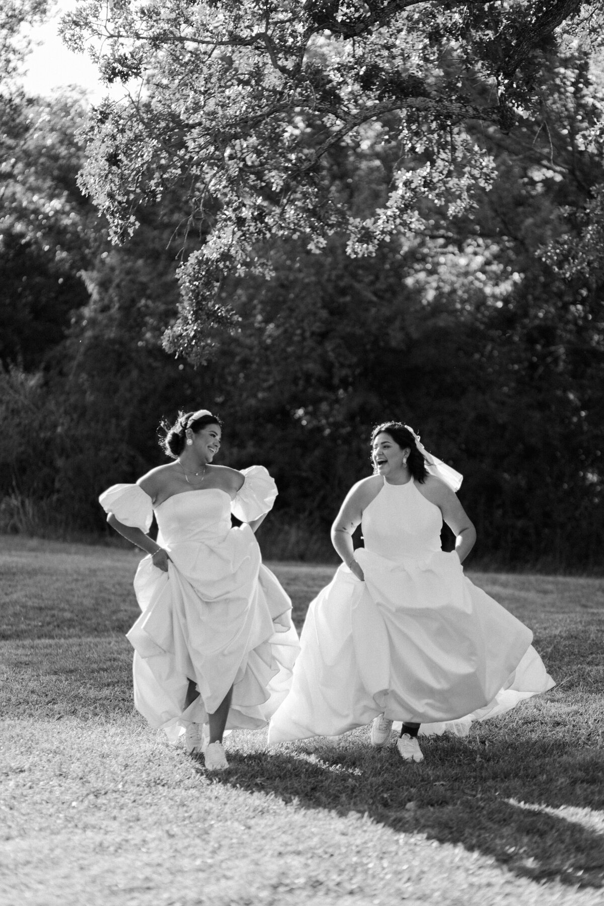Black and white portrait of two brides playfully frolicking on the grass on their wedding day in Fort Worth, Texas. The bride on the left is wearing a long, flowing white dress with pouffy sleeves. The bride on the right is wearing a long, flowing, sleeveless dress with a veil.