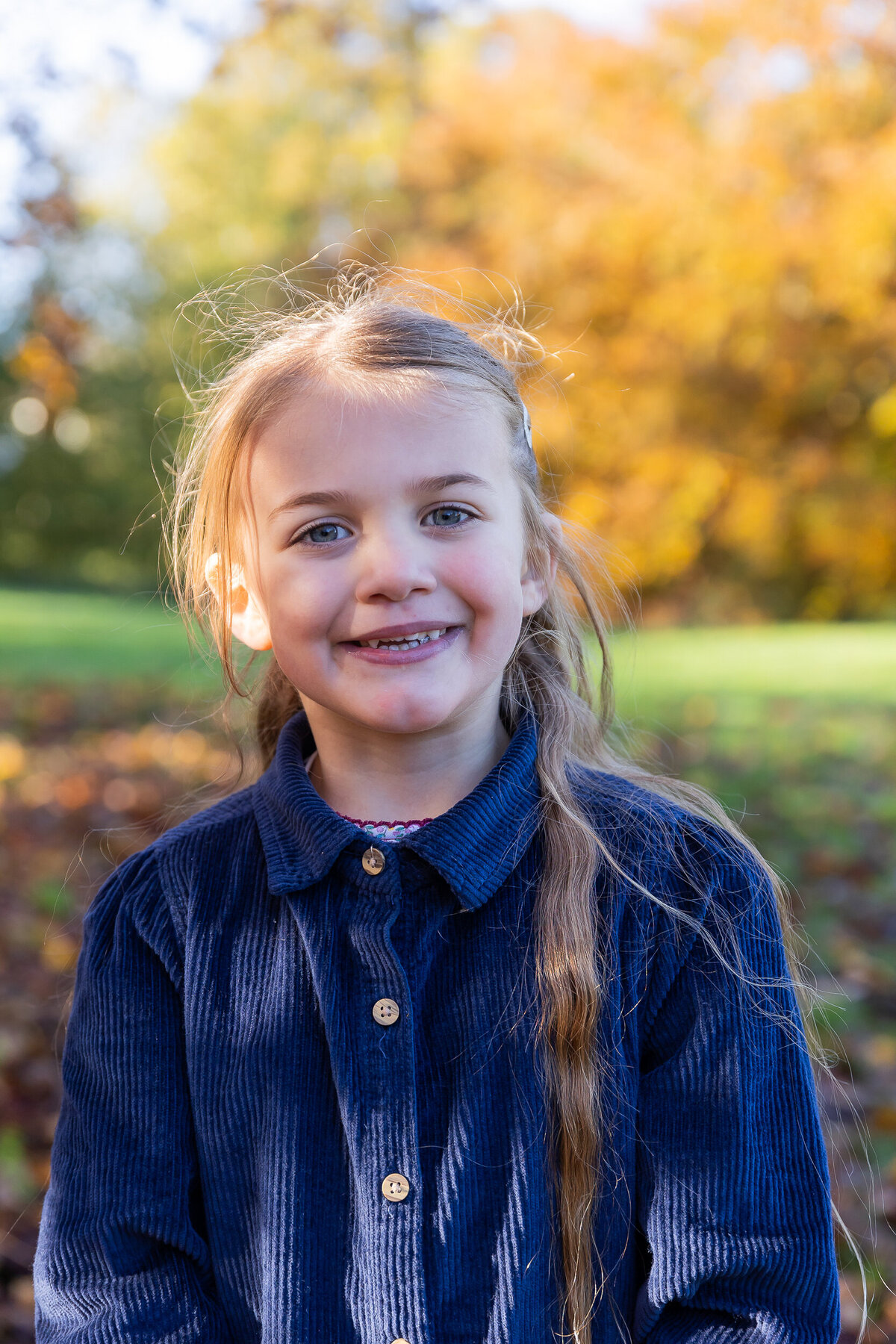 Young girl smiling in a park with autumn trees in the background. she wears a blue shirt and has braided hair.