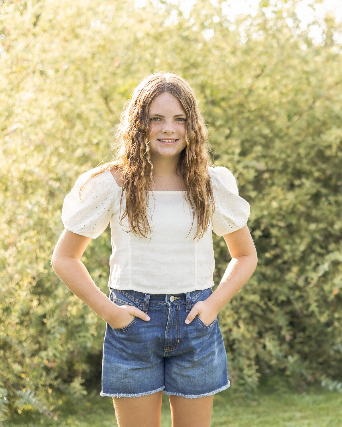 Older daughter standing in front of a bush in the grass, smiling at the camera with her hands in her pockets.  Older daughter is wearing blue jean shorts, a white short sleeve shirt, and has long brown hair. 
