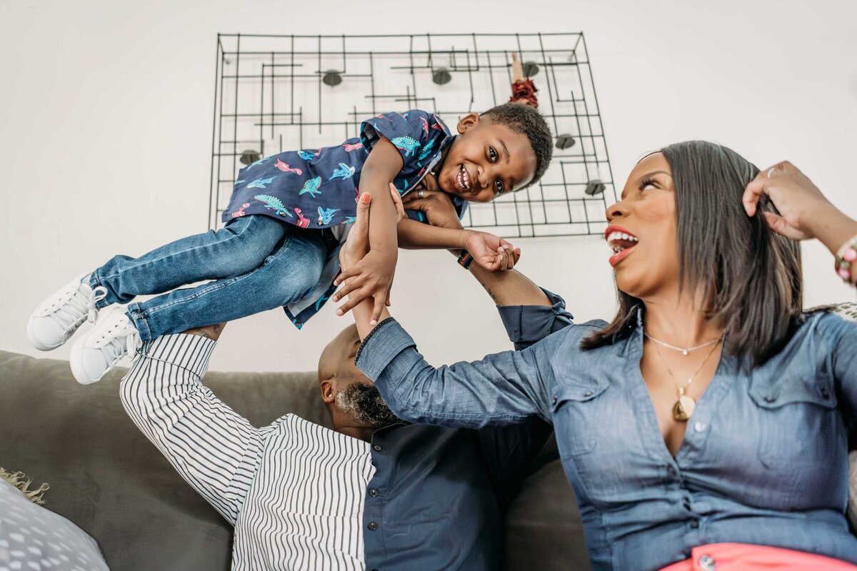 playful portrait of beautiful black family of three taken at upward angle of boy being held in air by dad with mom playfully watching