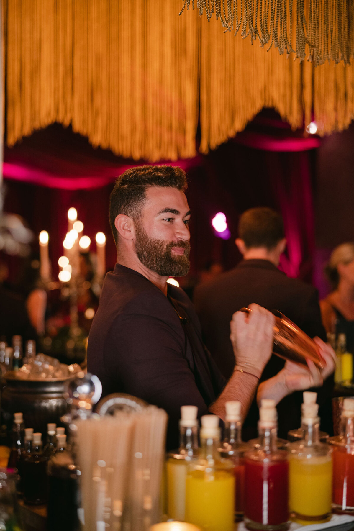 The photo captures a magical moment at a tented wedding in Virginia, expertly documented by the talented photographer Elisabetta Marzetti. In addition, Clayton Films provided videography services to capture the special moments of the event. In the midst of the festivities, a bearded man in a dark jacket skillfully shakes a cocktail shaker behind a beautifully decorated bar, featuring a variety of bottles and drink ingredients. The bar area exudes a cozy ambience with its warm lighting in red and golden hues, creating a perfect backdrop for the joyful occasion. In the softly focused background, other attendees can be seen, adding to the lively and celebratory atmosphere of the gathering.