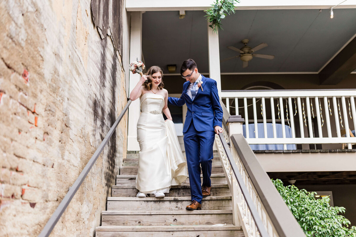 A bride in a white dress and a groom in a blue suit walk down wooden outdoor stairs together, holding hands and smiling, with a building and greenery in the background.