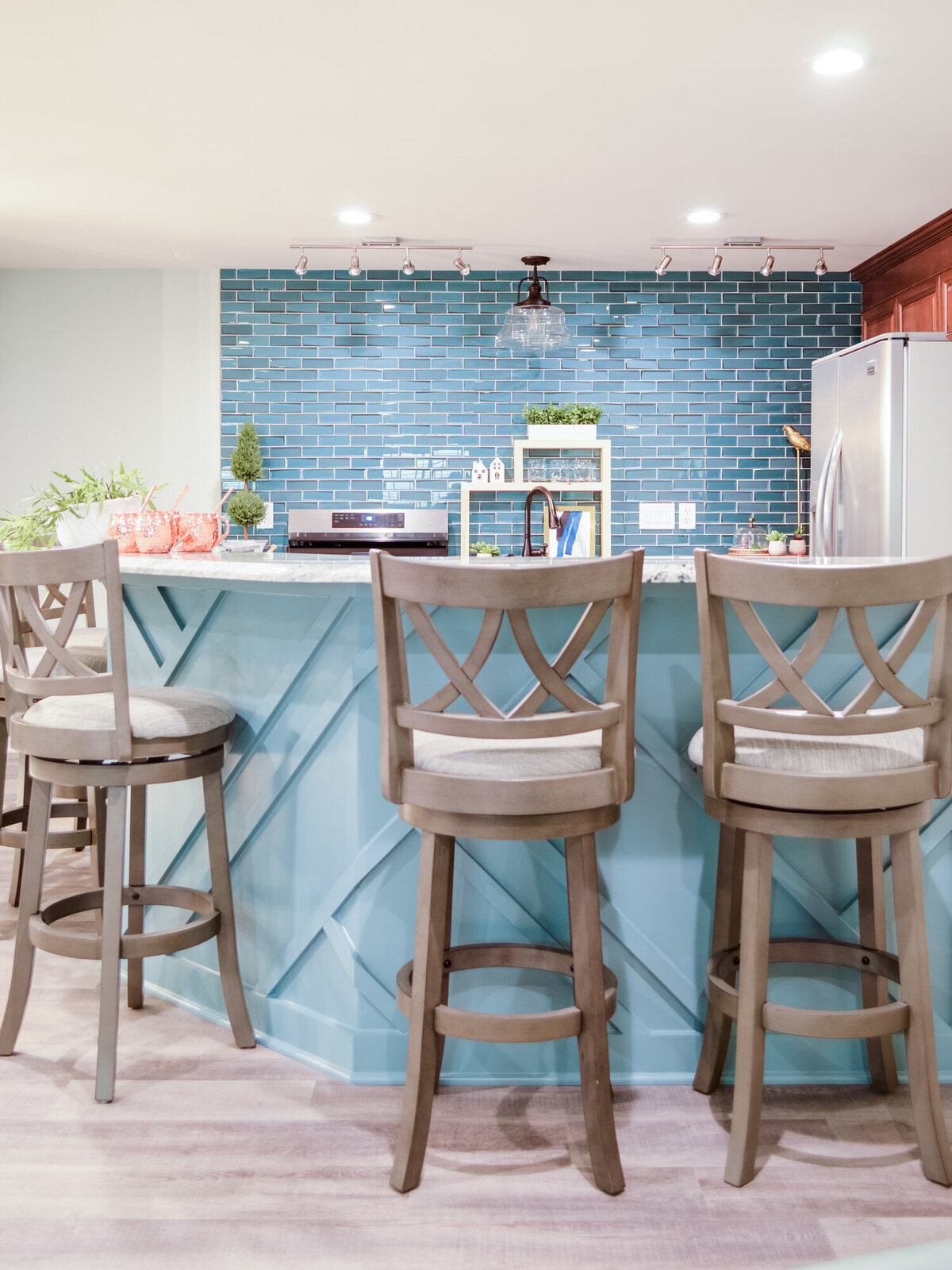 A well-lit kitchen island area featuring a light blue base with a geometric paneled design and a white countertop. Three Stools with light gray fabric seats and backs are positioned around the island. The stool in the center is angled slightly towards the viewer, showcasing its cross-back design. Behind the island, the wall is tiled with horizontal rows of glossy light blue subway tiles. A stainless steel microwave is built into the island. A small white shelf unit with greenery and other decorative items sits on the countertop. A stainless steel refrigerator is visible in the background on the right. Track lighting illuminates the backsplash, and recessed lighting is in the ceiling. The floor is covered with light-colored wood-look planks.