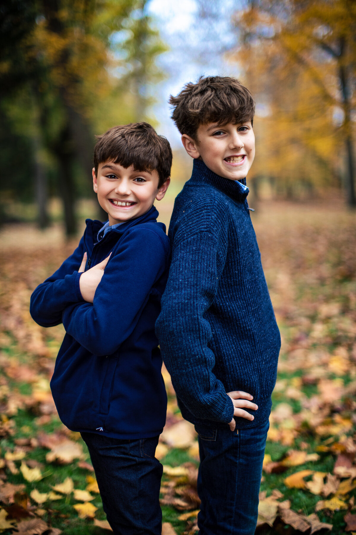 Brothers pose back to back wearing blue sweaters with orange fall leaves covering the ground around them during the children's mini session near Pittsburgh Pennsylvania