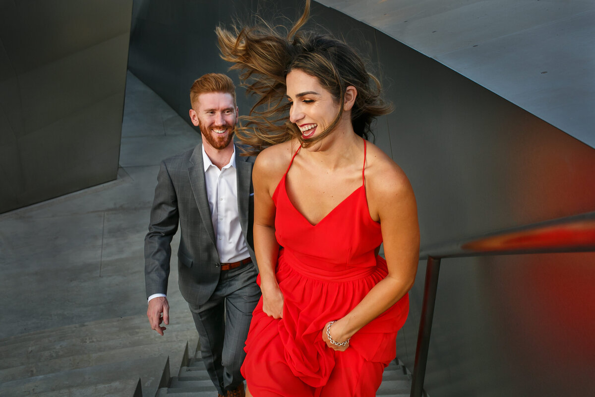 A soon to be bride and her fiance run up the stairs as she wears a striking red dress at Walt Disney Concert Hall in Los Angeles, photo by Tanya Parada
