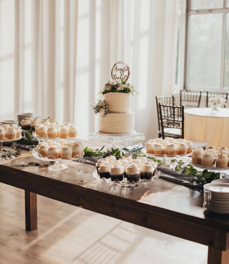 A white textured two tier wedding cake sits on a wooden table surrounded by clear stands of different flavors of cupcakes.