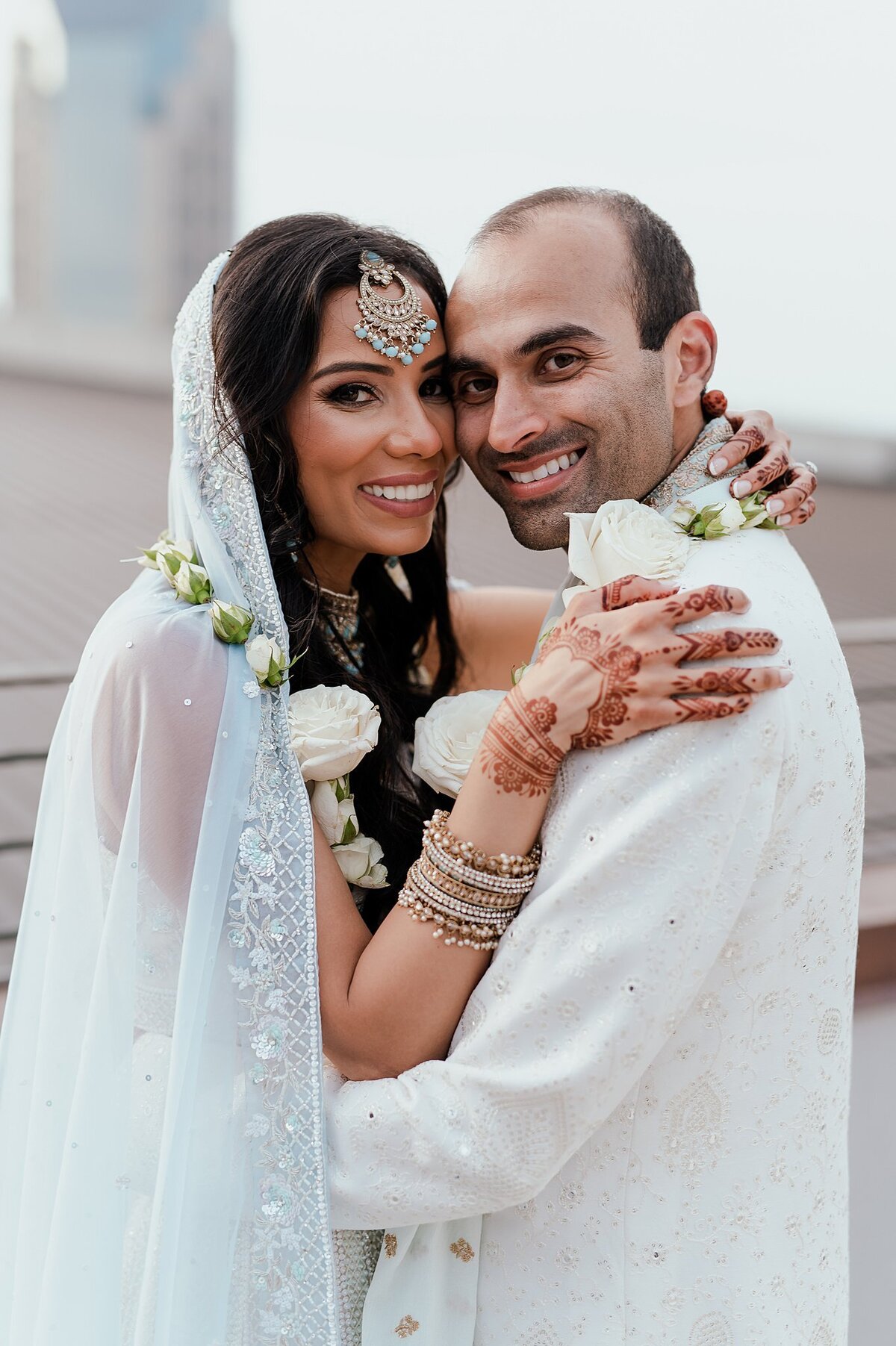 Indian bride with wedding henna mendhi on her hands smiles at the camera. The Hindu bride is wearing a light blue dupatta shawl with light blue and gold Indian wedding jewelry and a white floral varmala garland. The Hindu groom is wearing a white sherwani with a white floral varmala garland and is smiling at the camera for their Nashville Indian Wedding.