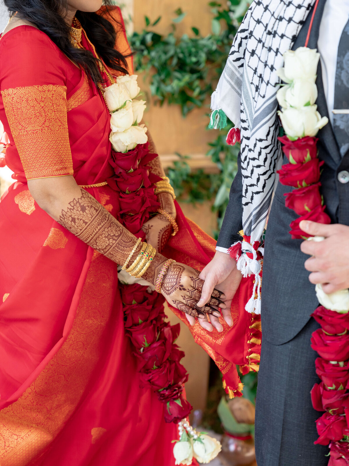 A couple holds hands during a wedding ceremony. The bride wears a red sari with intricate henna on her hands, while the groom wears a suit. Both have garlands of red and white roses. Green foliage decorates the background.