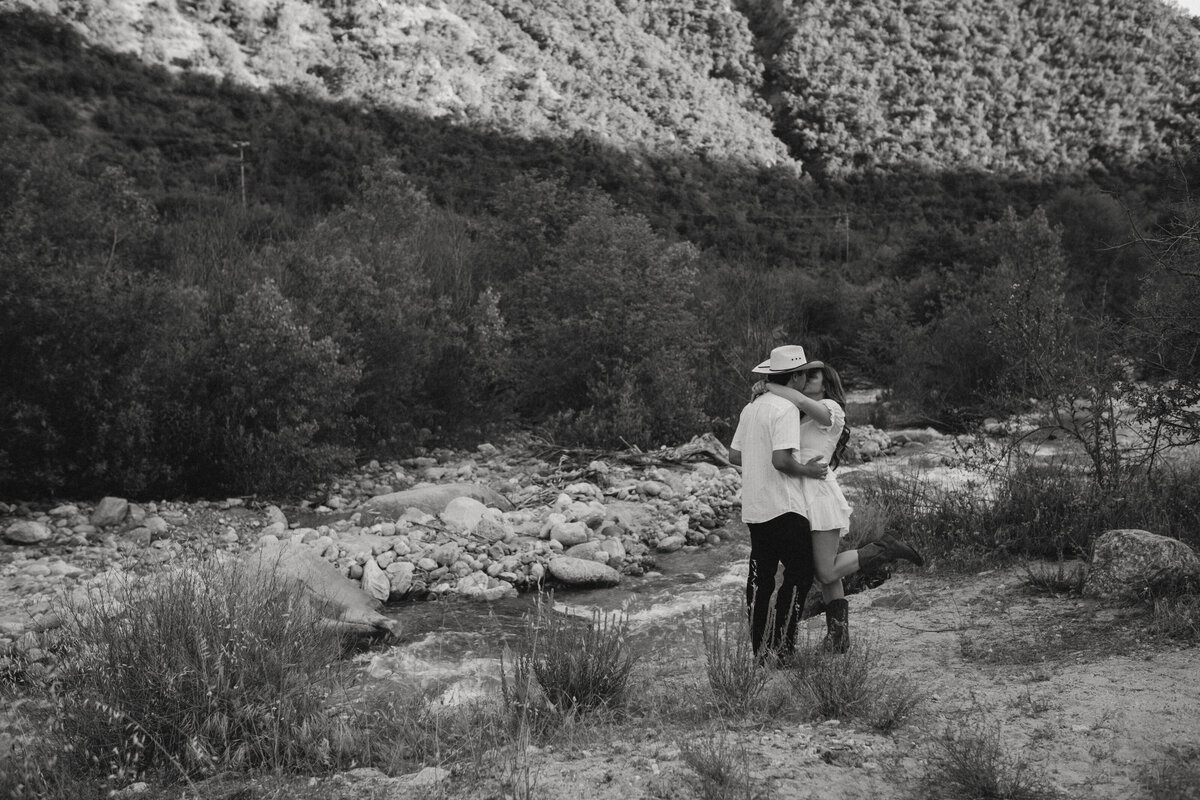 black and white couples photo kissing in front of river with mountain's in the back round