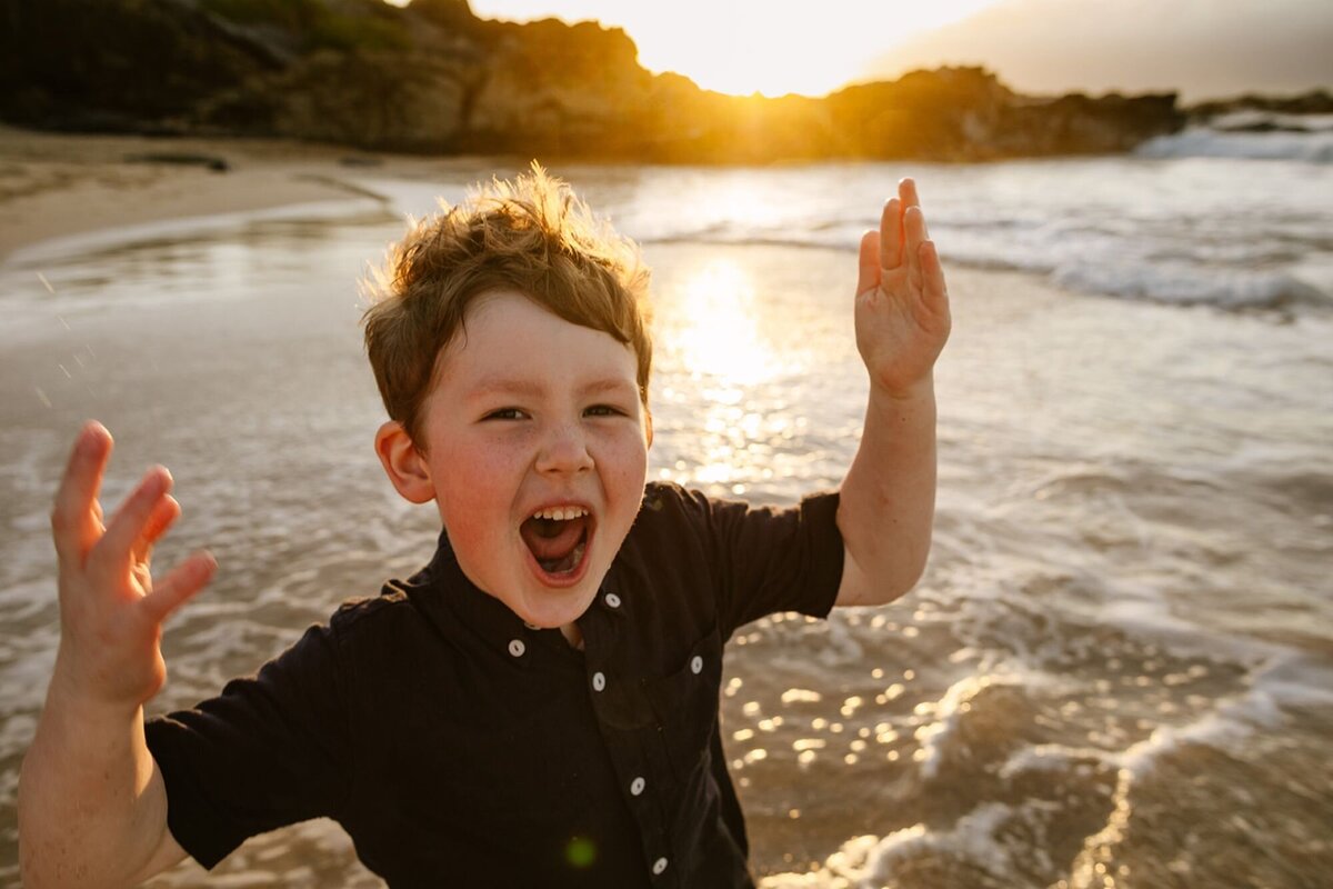 A little boy throws his arms up in the air, excited playing on the beach of Maui.