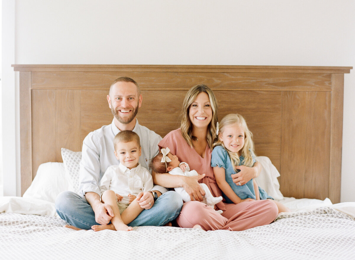 Family of 5 smiles at the camera while sitting on their bed during a Raleigh NC newborn photography session. Photographed by newborn photographer Raleigh A.J. Dunlap Photography.
