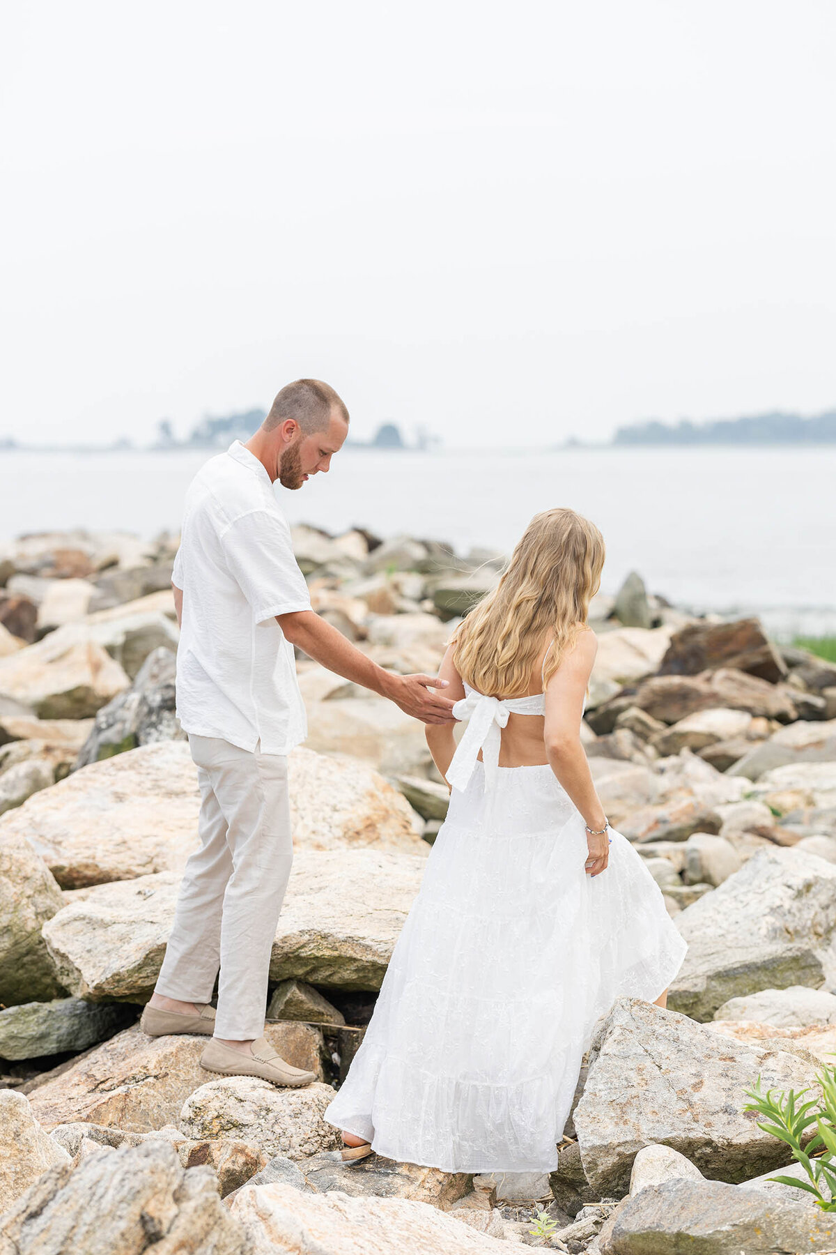 Couple walking along a rocky shoreline during a romantic Coastal Connecticut Session, woman in a white dress leading the way.
