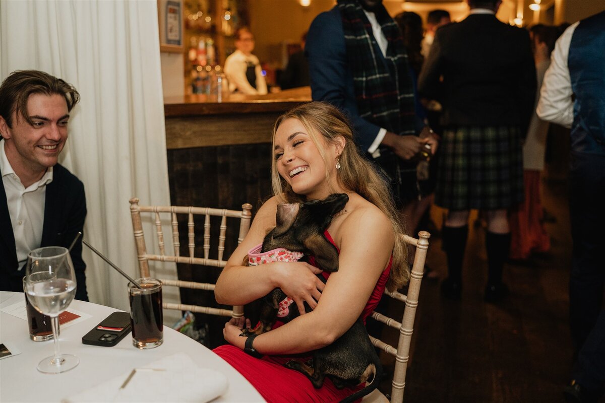 A woman holds a small dog and sits at a large round table during an evening wedding reception.