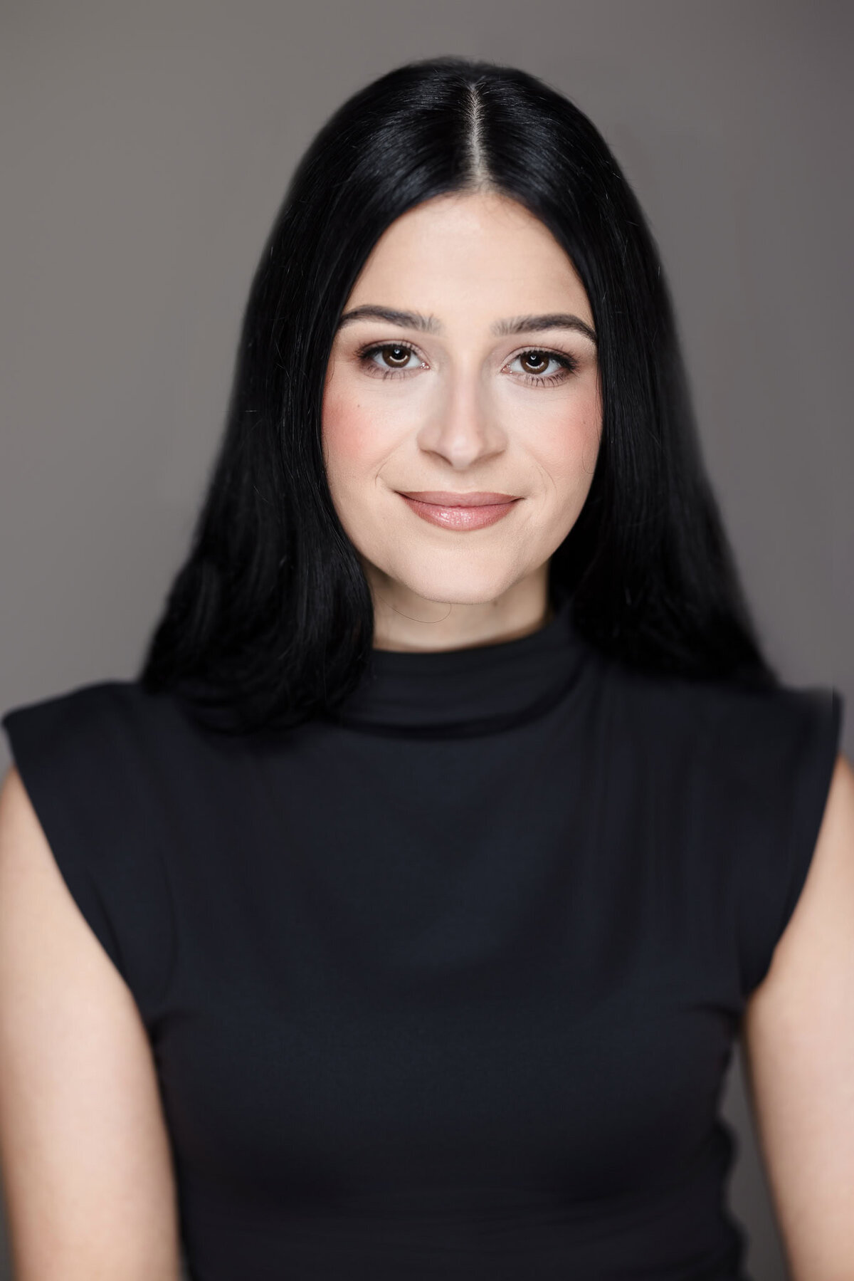 Corporate headshot of a woman with long black hair in a sleeveless black top, photographed against a gray background in Long Island.
