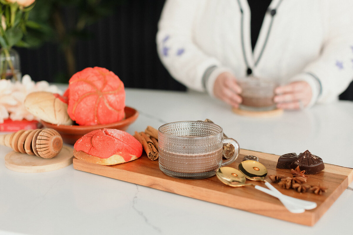 cClose up photo of items to make champurrado on a wood tray with woman's hands in background holding  cup of champurrado