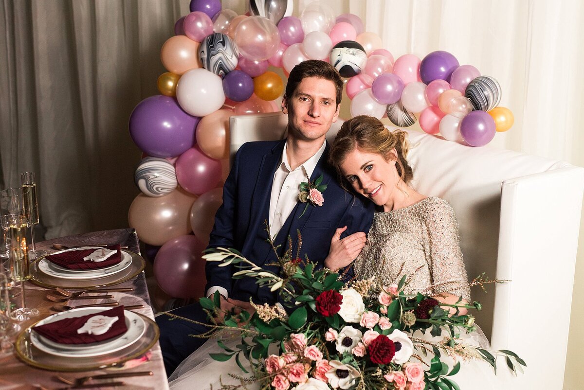 Bride and groom sit on a white leather loveseat decorated in with a purple, peach, gold, white, pink and marbled balloon garland. The bride, wearing a sequined wedding gown, is holding a large bouquet of red carnations, white anemone, pink roses and greenery. The sweetheart table is set against ivory drapery. The table is set with a swirling peach, purple, gold and ivory table cloth. The ivory plates rimmed with gold are set on gold chargers and covered with maroon napkins. On top of each napkin is a marble tile seating card.