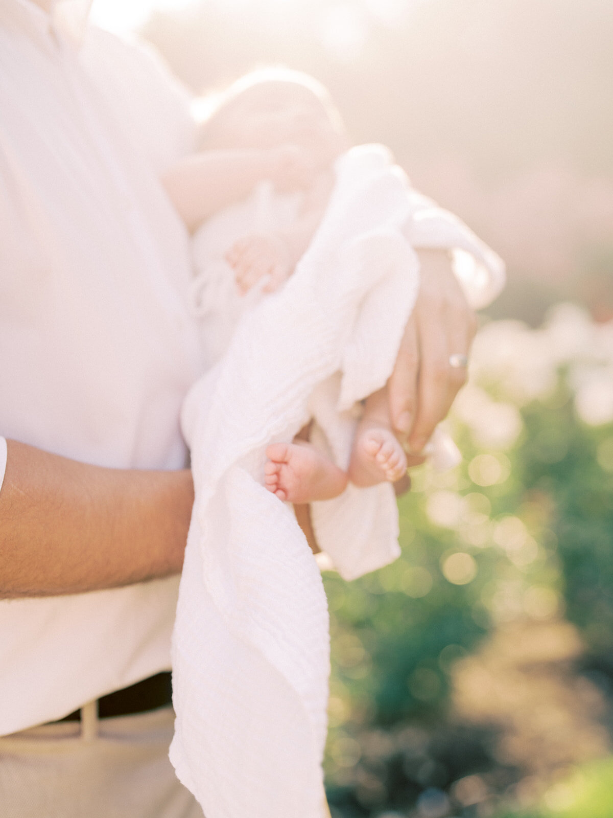 Two small baby feet stick out of a swaddle as the baby is held by her father outdoor.