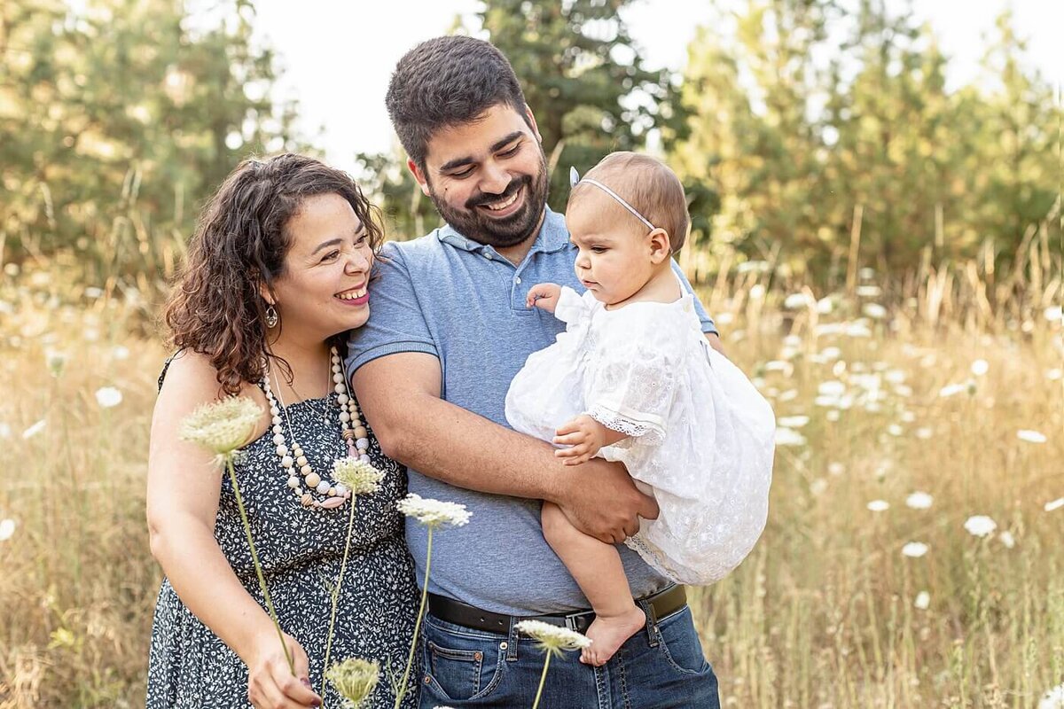 Family of three. Mom, Dad and baby girl dressed in blues and whites. Standing in a field of tall grasses and wildflowers. Mom and Dad are both looking at baby and smiling. Outdoor Portland Family Photography Session in the summer.