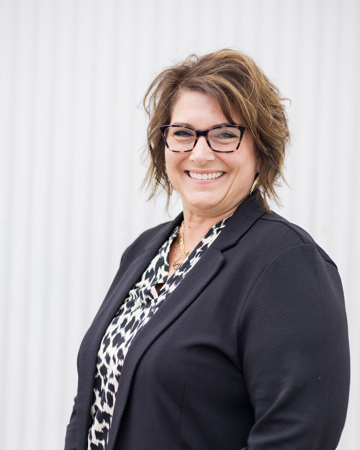 Woman standing in front of a white building with brunette blonde hair, wearing a black dress jacket with a white and black cheetah print shirt, wearing glasses, turned to the side, smiling at the camera