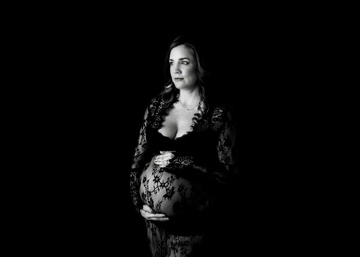 A pregnant woman in a black lace lingerie set stands in a studio holding her bump