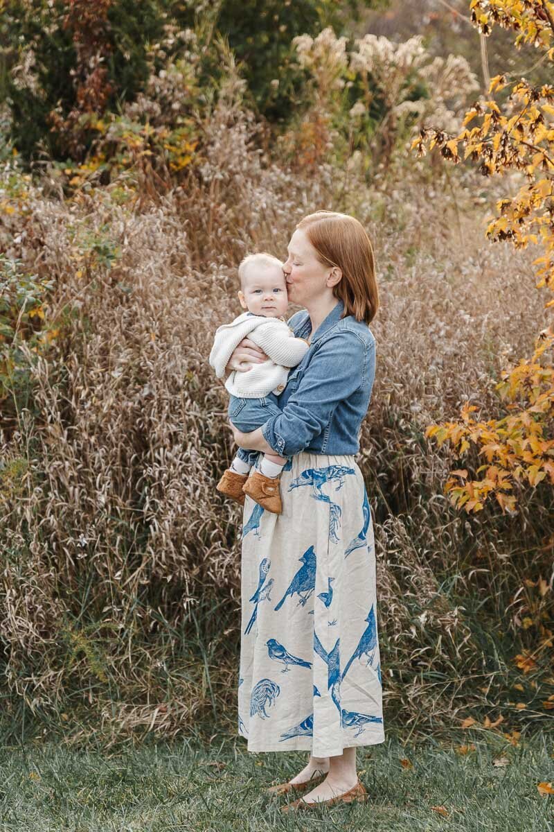 Show thumbnail preview A woman in a denim shirt and patterned skirt stands in a grassy area holding a baby, captured beautifully by a family photographer in Toronto. The background features tall grass and vibrant autumn trees.