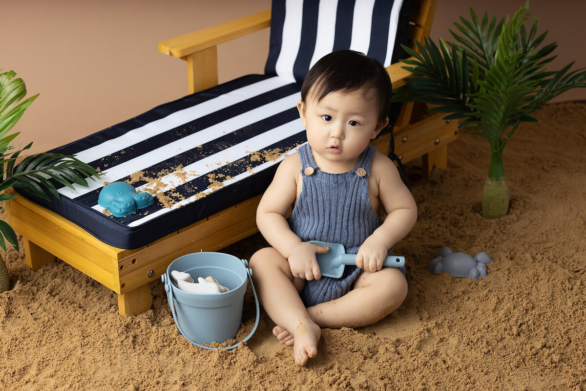 A baby boy in a striped romper plays with a shovel on a sandbox, surrounded by beach toys.