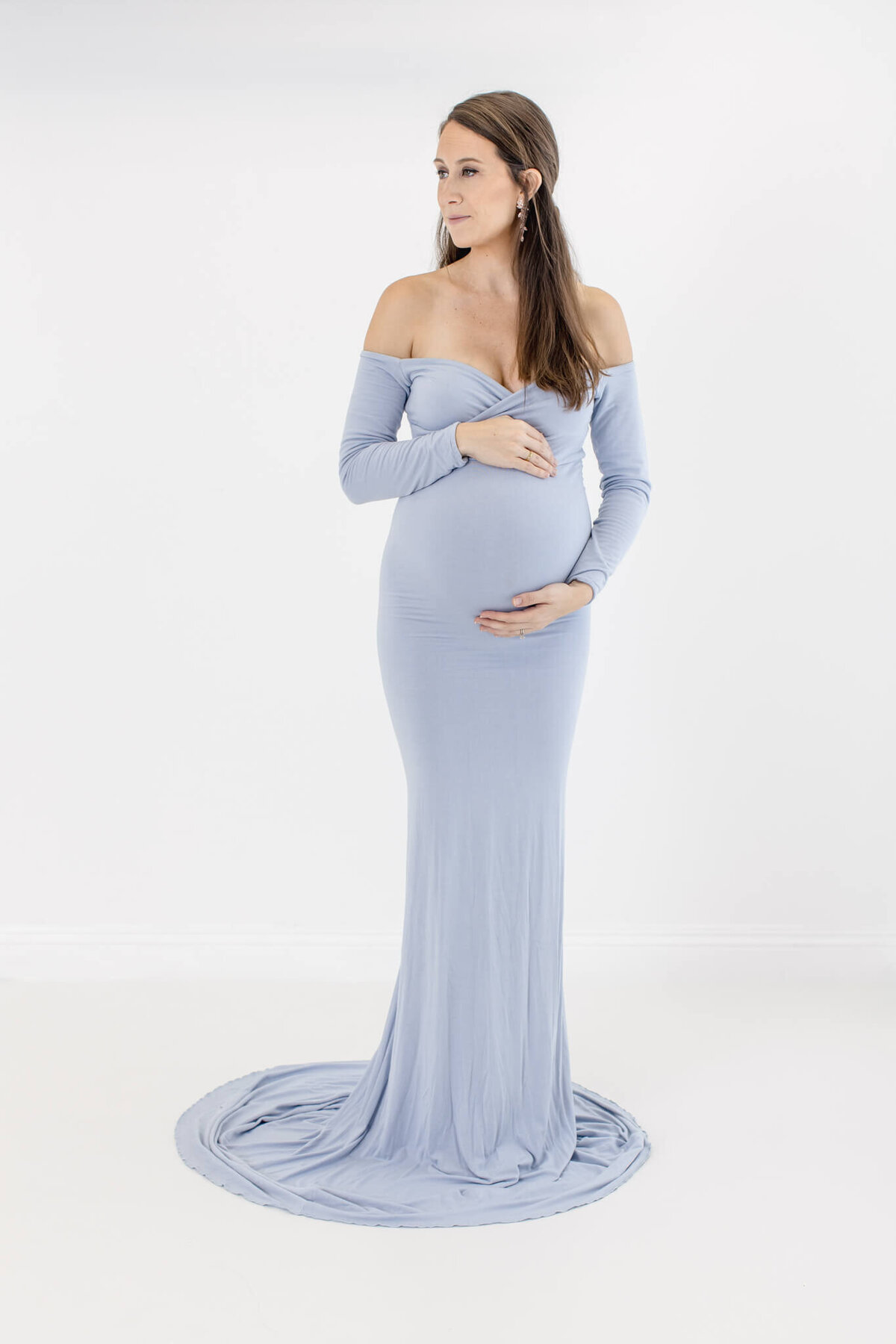 A pregnant woman in a form-fitting, long-sleeved, light blue gown gently cradles her belly. She stands against a plain white background, gazing to her right. The dress has a small train pooling on the floor.