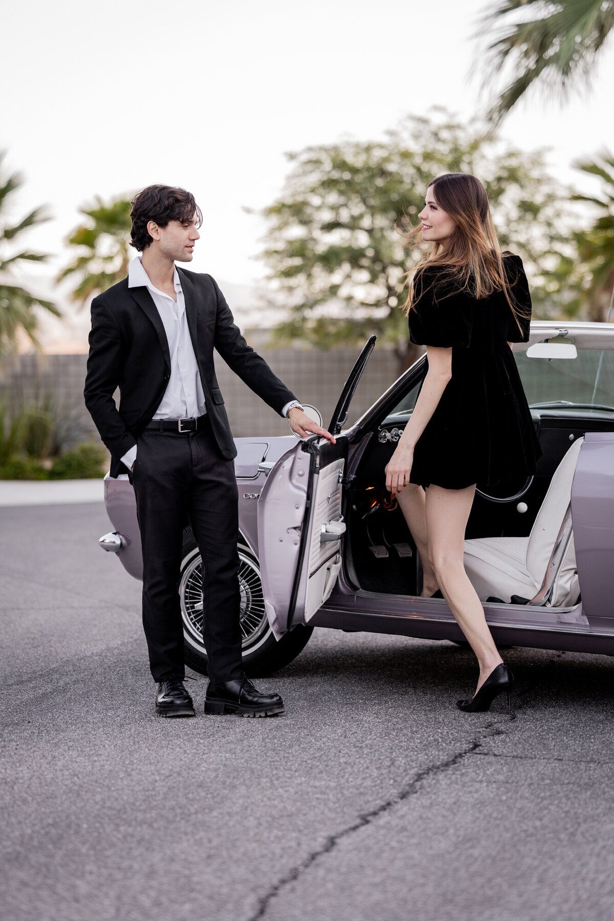 One partner opens the car door as the other steps out in a black dress and heels on a Palm Springs road lined with palms.